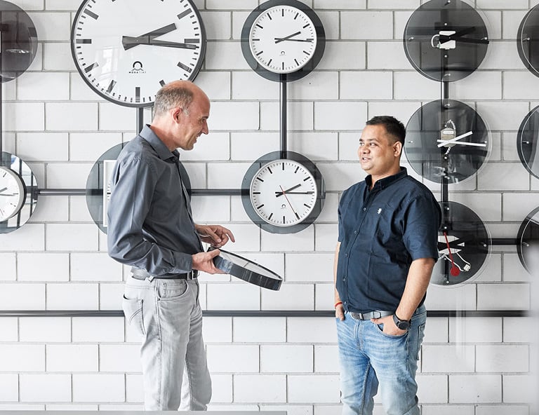 Two people are standing in front of a wall with various MOBATIME watch models and discussing the topic of GNSS.
