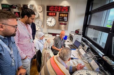 Ingenieros y representantes del proyecto observan las pruebas en vivo de los relojes maestro y esclavo durante la Prueba de Aceptación en Fábrica (FAT) para el proyecto de Tren de Alta Velocidad de Egipto en la sede de Moser-Baer, con las pantallas horarias sincronizadas visibles al fondo.